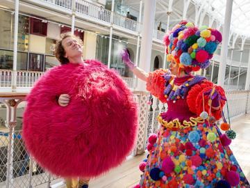 Photo of 2 people. One dress with a bright pink fluffy dress and the other in a dress made from coloured pom poms