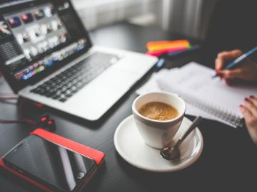 Person sitting at desk with notebook, pens, mobile, laptop and cup of coffee