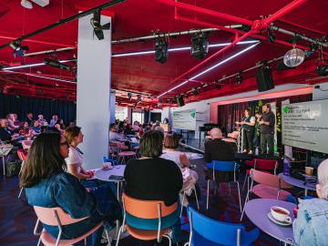 Lots of people sat on colourful chairs whilst someone stands on a stage and talks to them