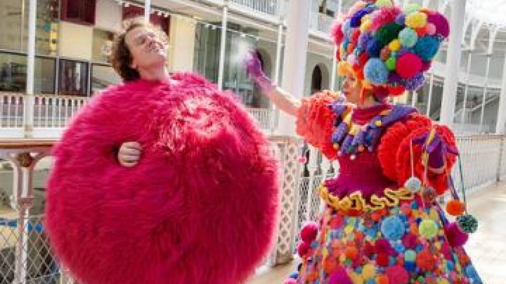 Photo of 2 people. One dress with a bright pink fluffy dress and the other in a dress made from coloured pom poms