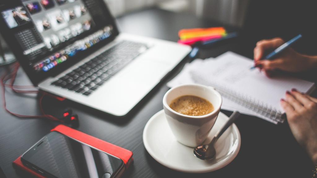 Person sitting at desk with notebook, pens, mobile, laptop and cup of coffee